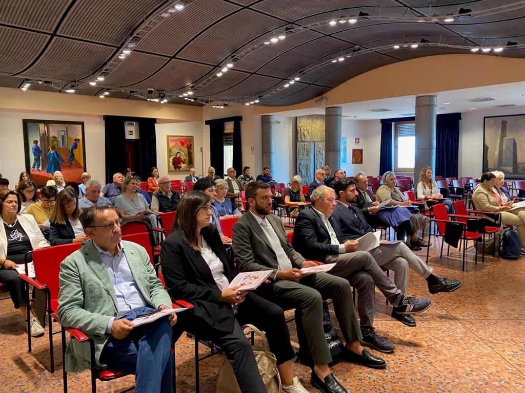 Un gruppo di persone partecipano a un incontro in una sala, con un'attenzione rivolta verso relatori e presentazioni, circondati da opere d'arte sulle pareti.