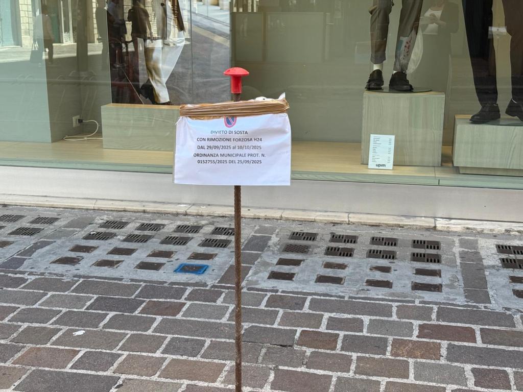 A sign posted on a metal pole indicating a parking prohibition with details about enforcement dates, located in front of a store showcasing mannequins in window.