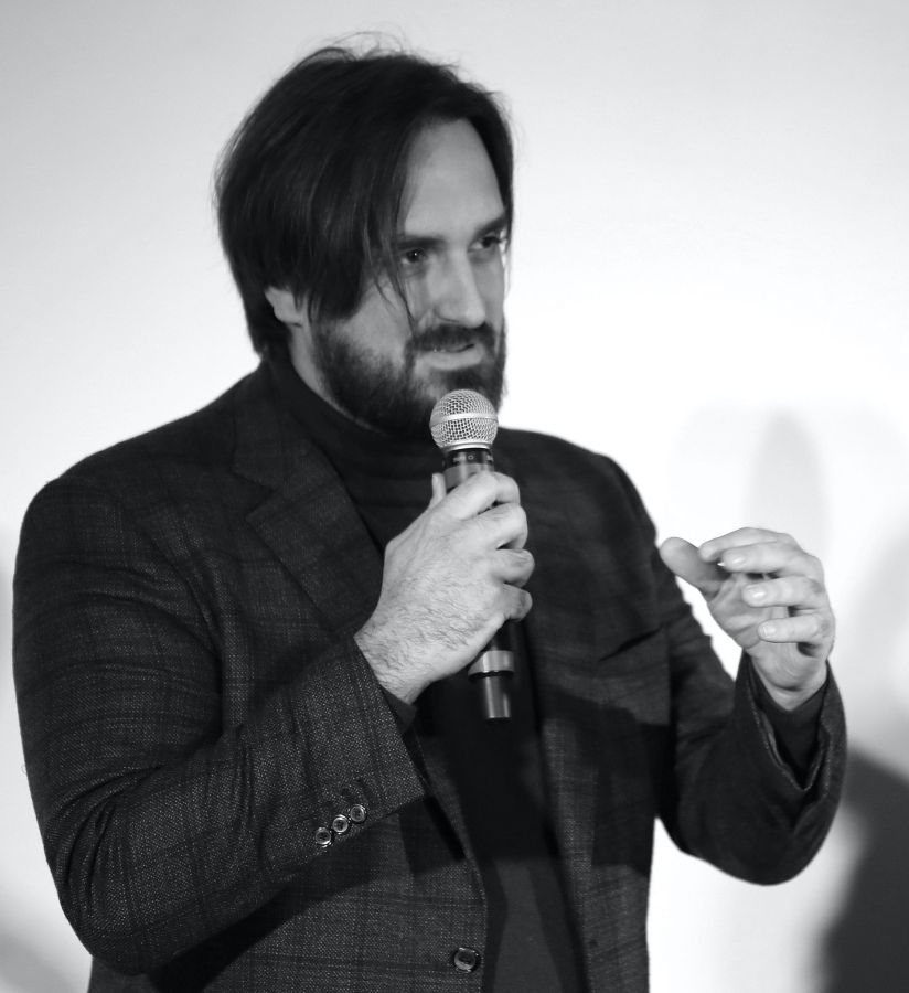 Un uomo con una giacca scura tiene un microfono mentre parla, gesticolando con la mano. Lo sfondo è neutro e sfocato.