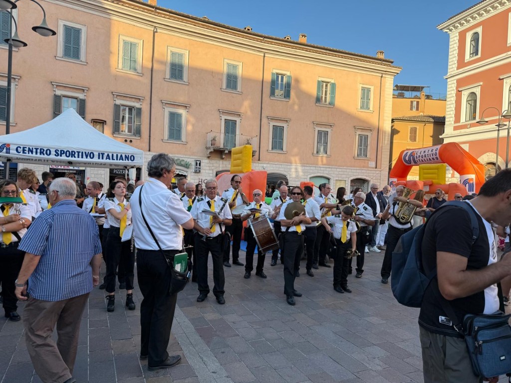 A band dressed in white shirts and yellow ties performs in a town square, with a large inflatable structure in the background and spectators gathered around.