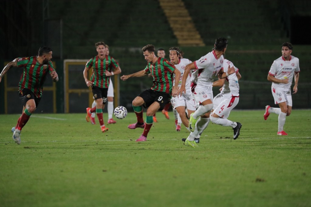 A soccer match in progress, featuring players in green and red striped jerseys and white jerseys. A player wearing number 9 in a striped kit is attempting to control the ball while being pursued by opposing players.