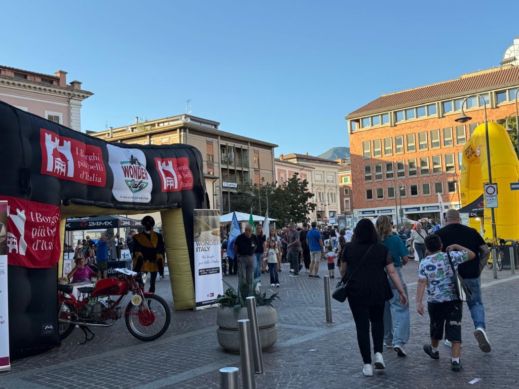A lively street scene during an event, featuring an inflatable archway with banners promoting 'Wonderful Italy' and 'The Most Beautiful Villages of Italy'. People are walking around, enjoying the festivities, with stalls visible in the background.