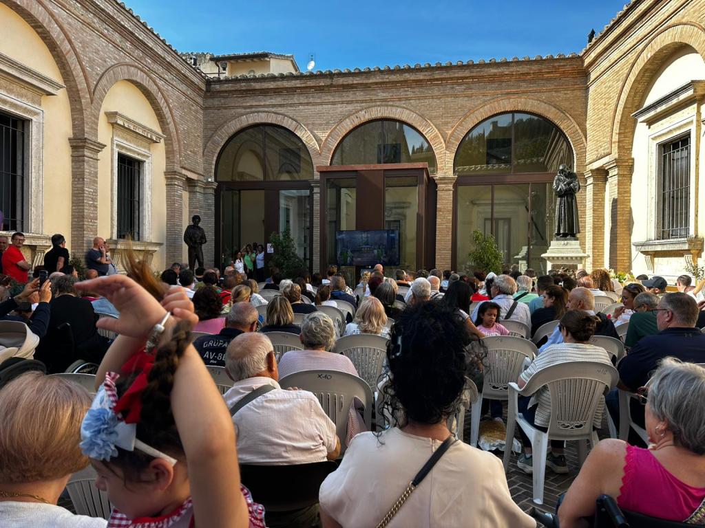 A large crowd of people seated outdoors in a courtyard, watching a screen showing a live ceremony. Some individuals are raising their hands, with a statue visible in the background.