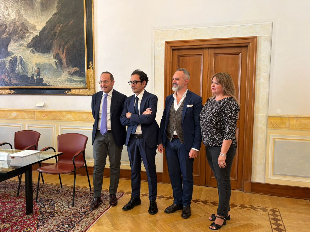 Gruppo di quattro persone posizionati accanto a un tavolo in una sala con decorazioni storiche e un dipinto sullo sfondo, sorridendo per un evento di beneficenza.