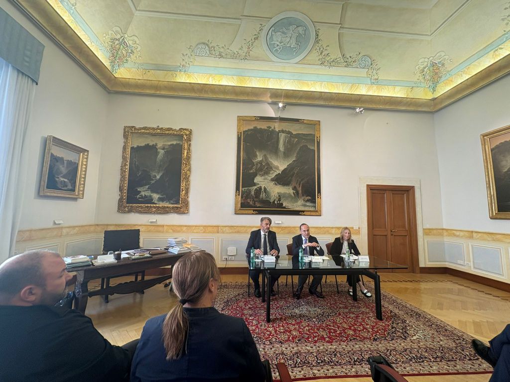 A formal meeting taking place in a historic room with paintings on the walls. Three individuals are seated at a table, engaged in discussion, while attendees listen from the background.