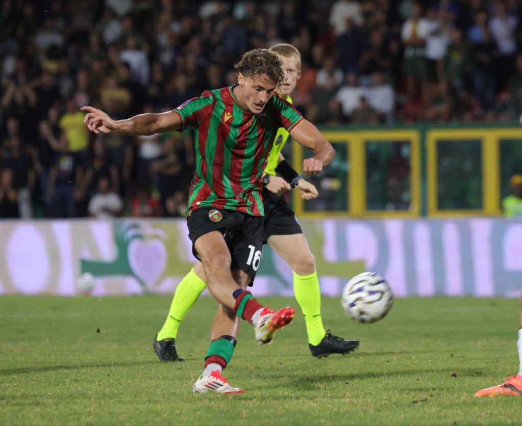 Un giocatore di calcio in maglia a strisce verdi e rosse sta calciando un pallone in un campo affollato durante una partita.