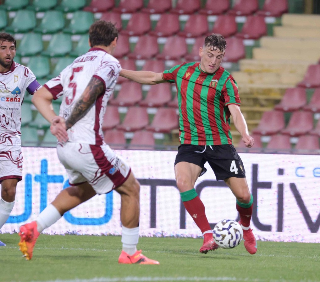 Un giocatore in maglia rossa e verde cerca di controllare il pallone mentre un avversario in maglia bianca lo ostacola durante una partita di calcio.