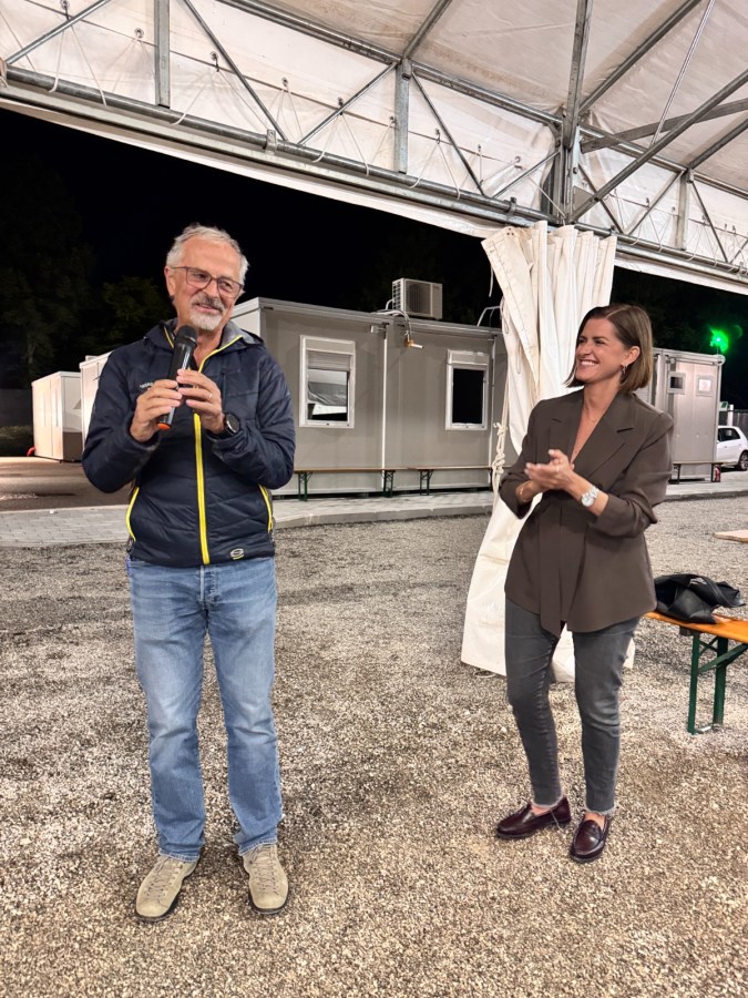 Un uomo con occhiali che parla al microfono, mentre una donna accanto applaude. Sullo sfondo si vedono moduli prefabbricati in un'area allestita per un evento.