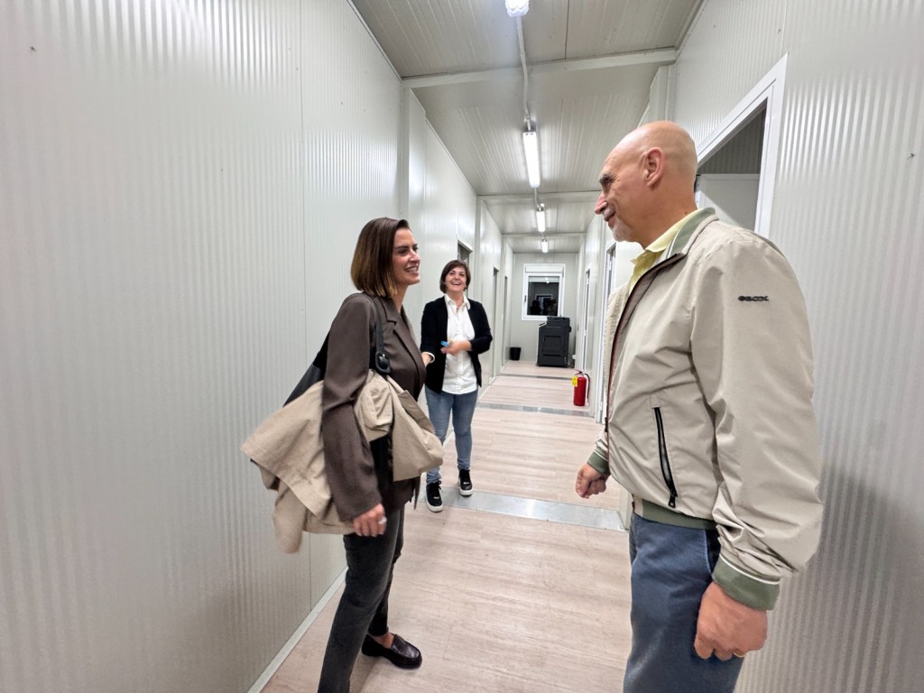 Tre persone conversano in un corridoio all'interno di un campo base, circondati da pareti in lamiera metallica. Due donne sorridono mentre un uomo ascolta, creando un'atmosfera amichevole e collaborativa.