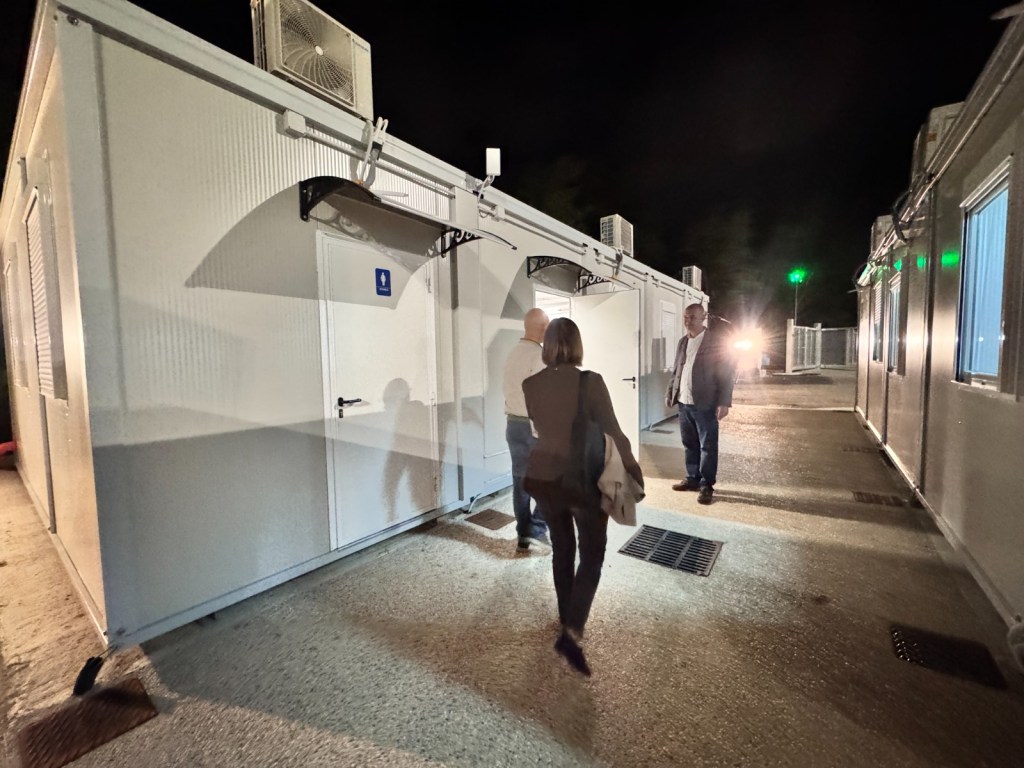 Persone che camminano verso moduli abitativi temporanei allestiti per il campo base del Metrobus di Perugia, con illuminazione notturna.