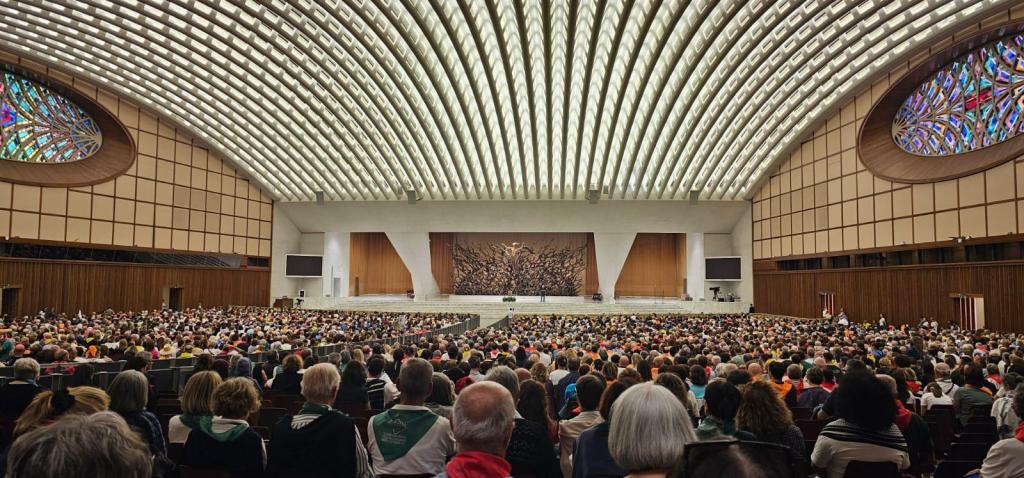 Interno dell'Aula Paolo VI in Vaticano, affollato da circa 6500 pellegrini umbri durante un evento religioso, con un grande arazzo sullo sfondo e vetrate colorate che illuminano la scena.