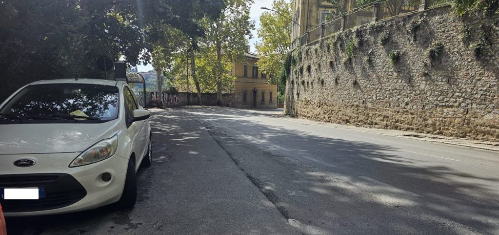 Un'auto bianca parcheggiata a lato di una strada asfaltata, con una parete di pietra e alberi sullo sfondo.