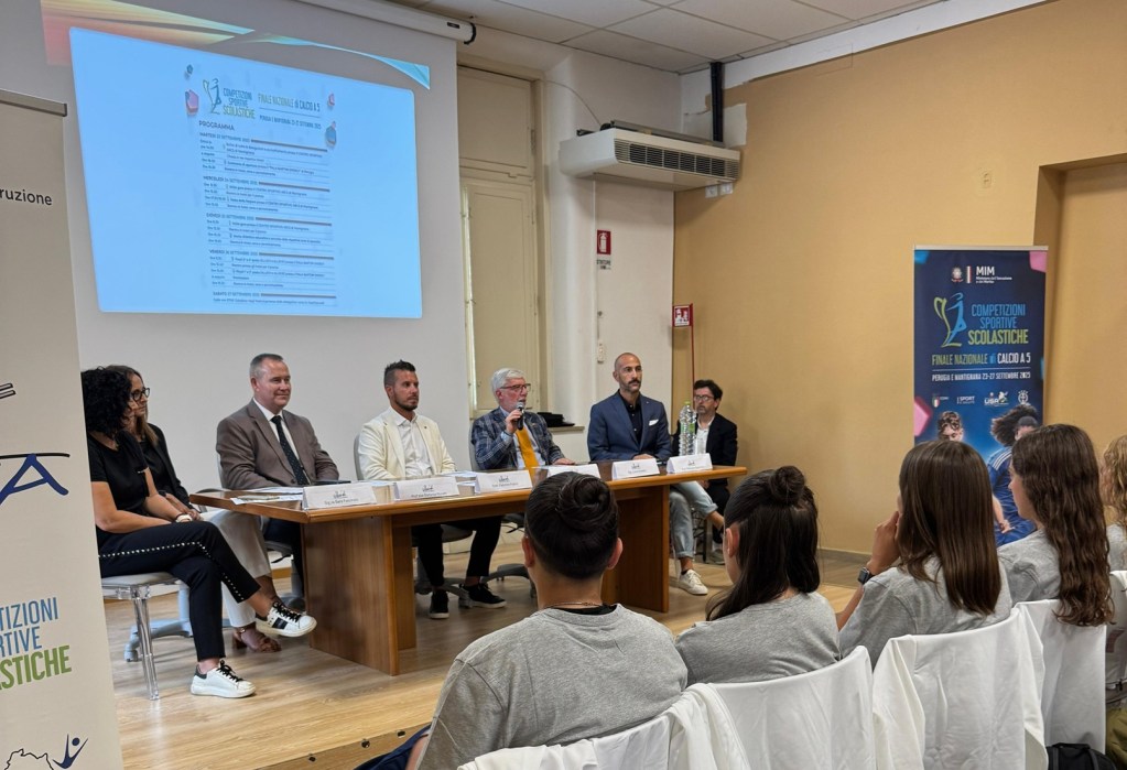 Una conferenza stampa per le finali nazionali di calcio a 5 scolastico, con relatori seduti a un tavolo e un grande schermo che mostra il programma dell'evento.