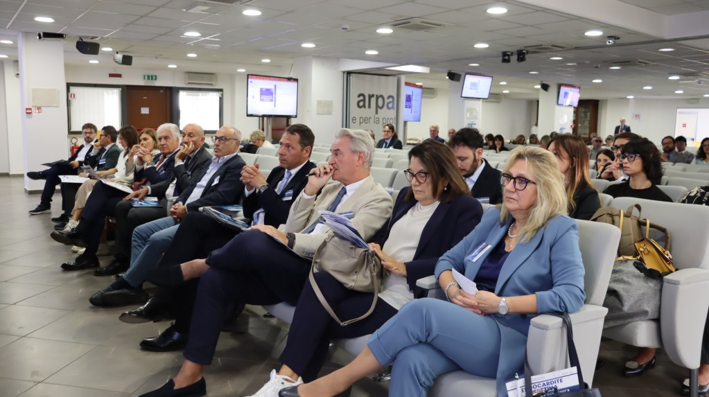 Una sala con diversi partecipanti seduti, coinvolti in una conferenza sulla endocardite infettiva, con schermi visibili sullo sfondo.