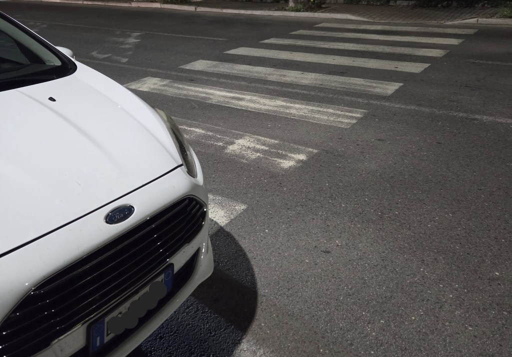 Auto bianca parcheggiata accanto a un passaggio pedonale usurato in una strada di Perugia, evidenziando la situazione di sosta irregolare.