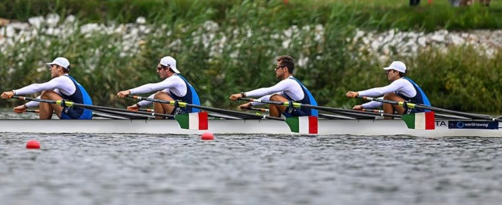A team of four rowers in a boat competing on a river, wearing matching uniforms with blue and white colors, featuring Italian flags on the boat.