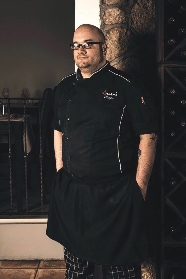 Chef Diego Mercuri in uniforme da lavoro, posando in un ristorante moderno con dettagli di arredo elegante e una cantina sullo sfondo.