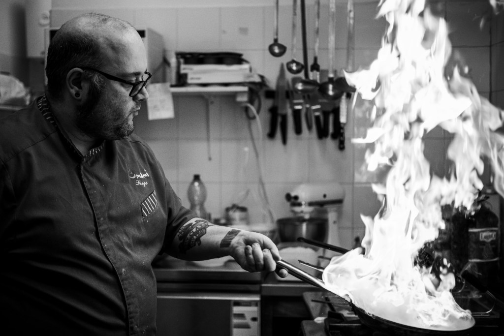 Un chef che cucina in una cucina professionale, mentre fiamme alte emergono da una padella.