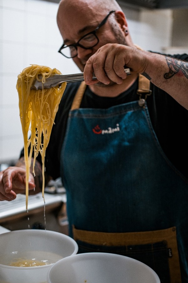 Uno chef mentre solleva spaghetti con delle pinze in una cucina professionale.