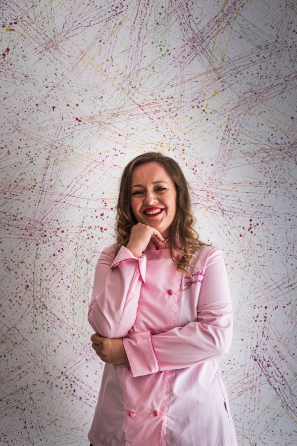 Chef sorridente in giacca rosa, posando con una mano sul mento davanti a uno sfondo colorato e astratto.