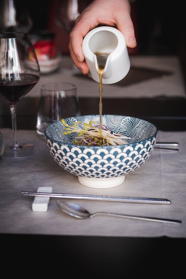 Una mano versa brodo caldo in una ciotola decorativa mentre un piatto di ramen con ingredienti freschi è posizionato sulla tavola. Si possono vedere un bicchiere di vino rosso e altri utensili sul tavolo.