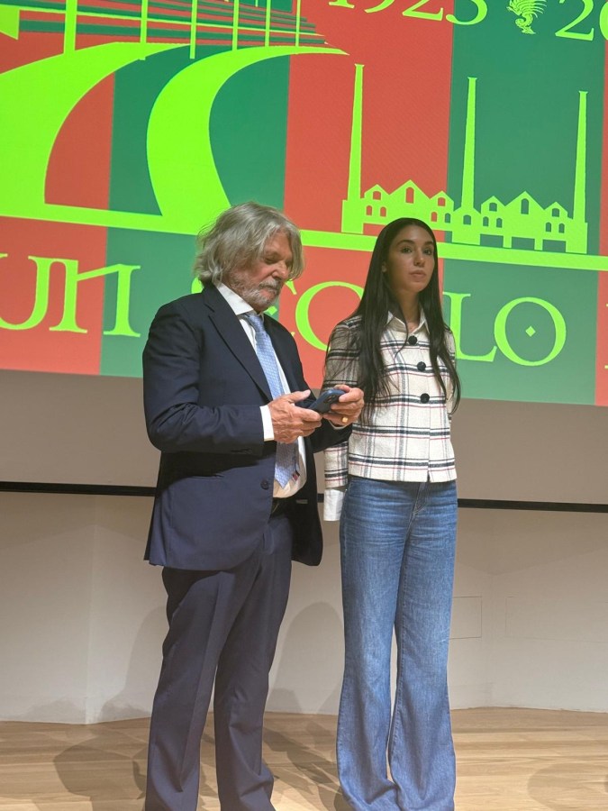 Due persone sul palco di una cerimonia, con uno schermo sullo sfondo che celebra il centenario della Ternana Calcio, indossano abbigliamento formale.