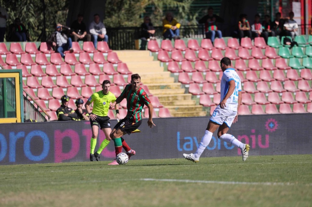 Giocatore in azione durante una partita di calcio, con pubblico sugli spalti e un poster pubblicitario visibile sullo sfondo.