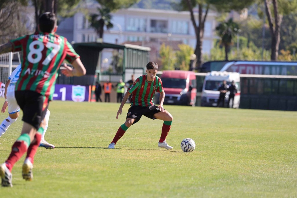Giocatore in azione durante una partita di calcio, indossando una maglietta a strisce rosse e verdi, con la palla vicino ai piedi e il campo sullo sfondo.