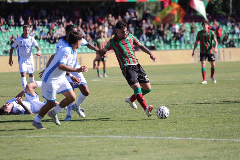 A football match scene featuring players in action on the field, with a focus on a player wearing a striped green and red jersey dribbling the ball while teammates and opponents are nearby.