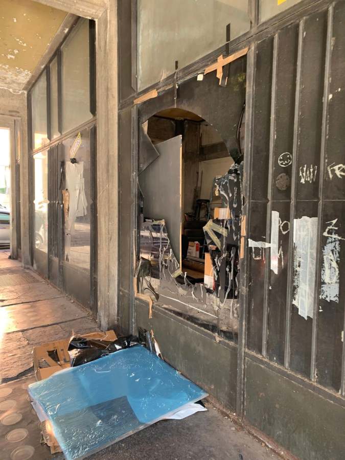 A broken glass window with fragments and debris lying on the ground outside a building, showing the interior through the damaged area.