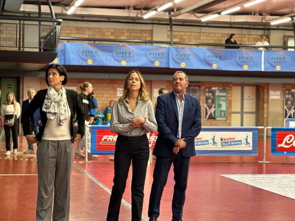 Tre persone in piedi all'interno di un palasport, osservando attentamente un evento di scherma.