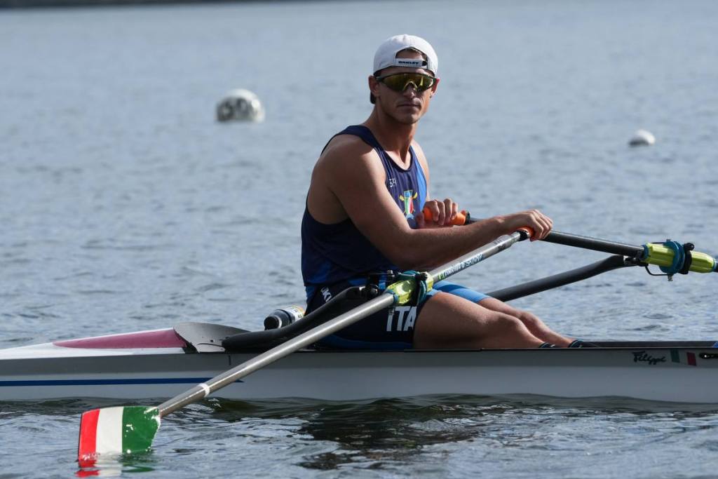 Un atleta italiano di canottaggio in competizione su un lago, vestito con una maglietta azzurra e pantaloni blu. Sulle spalle porta il distintivo dell'Italia. La barca ha un dettaglio con i colori della bandiera italiana.