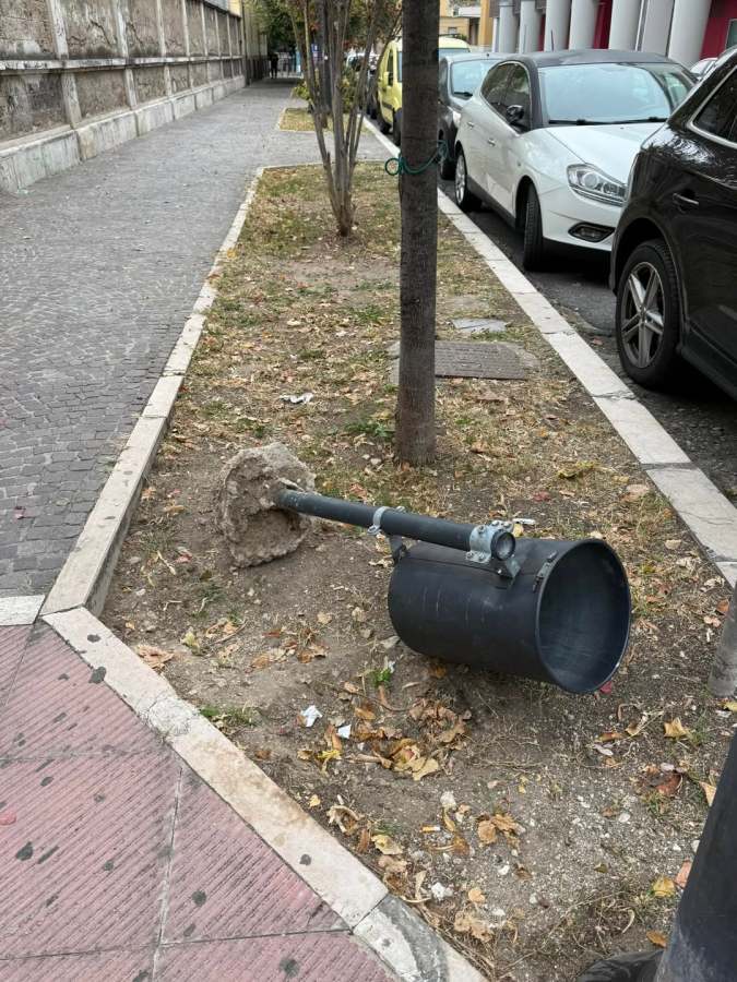 A black cylindrical container tipped over on a dirt patch next to a sidewalk, with fallen leaves surrounding it.