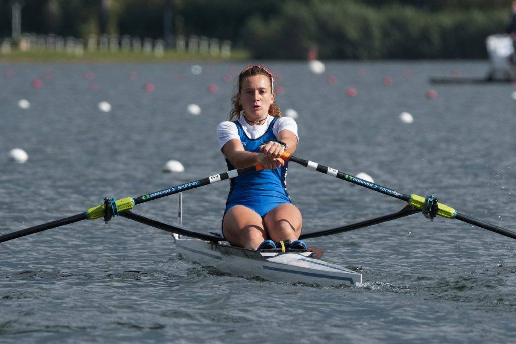Atleta che gareggia in una regata di canottaggio, remando in un singolo con un abbigliamento sportivo blu e bianco, su un lago con boe di gara visibili sullo sfondo.