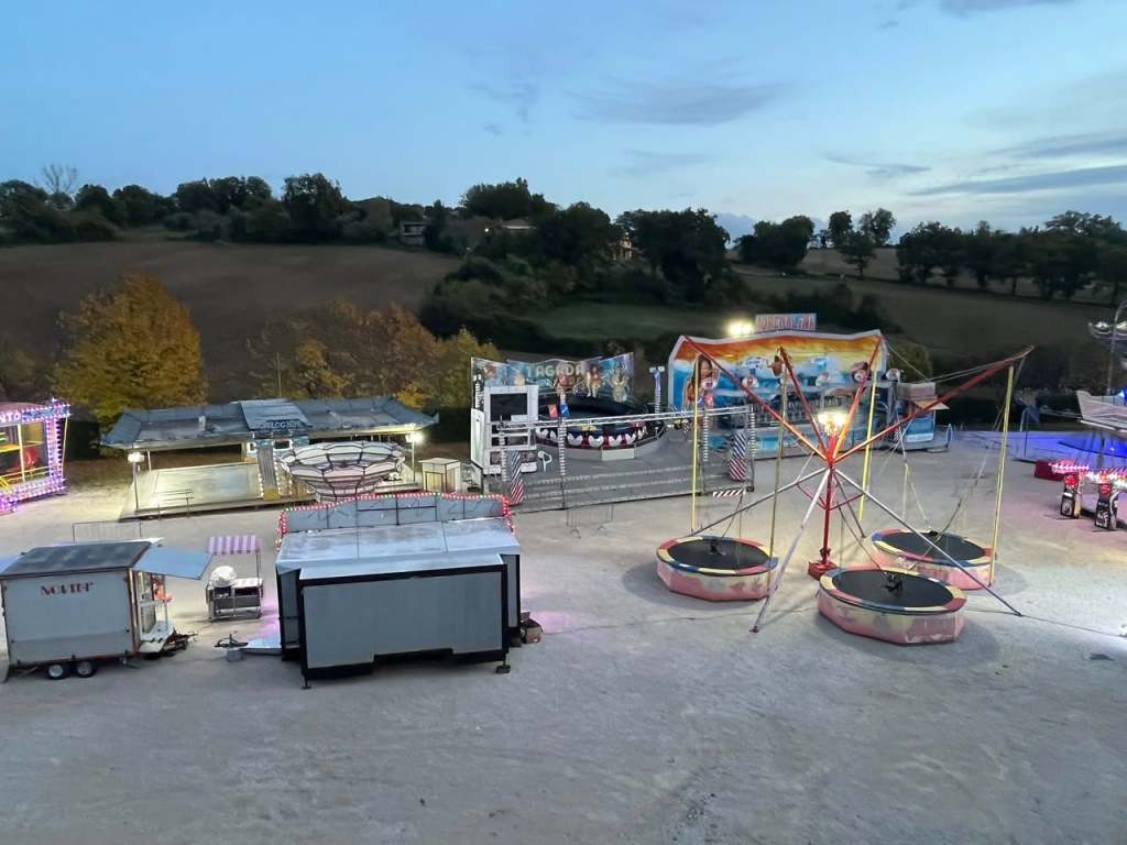 Vista panoramica di un luna park allestito, con giostre e attrazioni illuminate, nella campagna di Montecastrilli al tramonto.