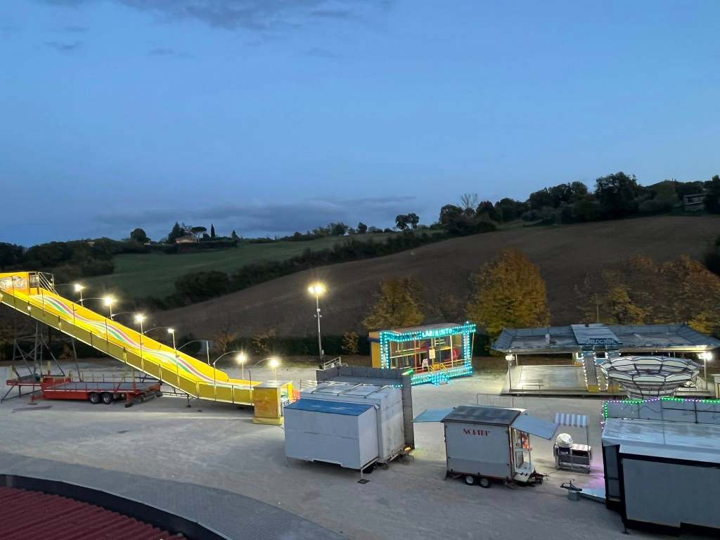 Panorama di un luna park allestito al centro fiere di Montecastrilli, con giostre illuminate e attrezzature in attesa di apertura. Sullo sfondo, colline verdi di autunno.