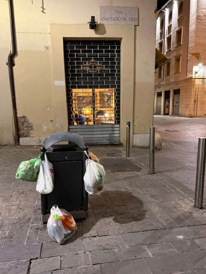 A trash can is filled with plastic bags of garbage, set against a backdrop of a dimly lit street and a locked storefront, featuring a sign that reads 'Via Anastasio De Filis'.