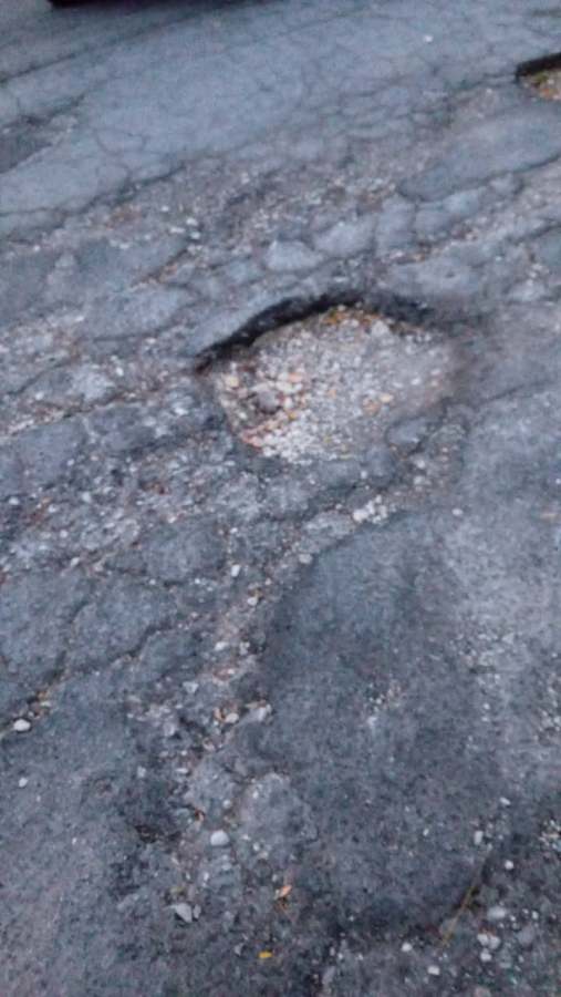 A close-up view of a damaged asphalt road showing a large pothole filled with loose gravel and debris.