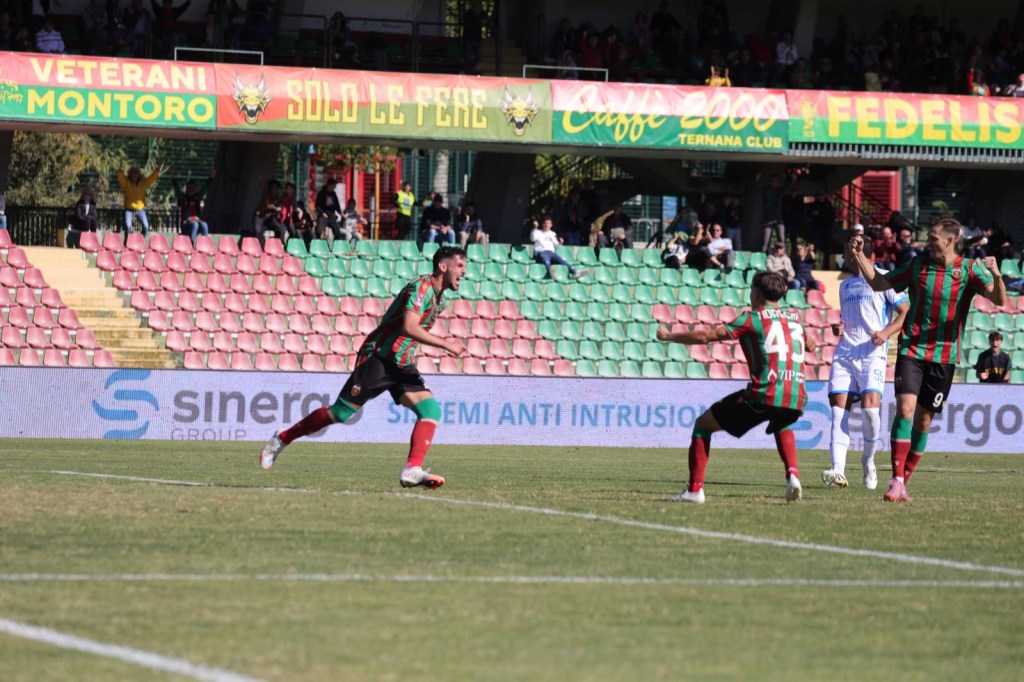 Giocatori della squadra Ternana esultano dopo un gol durante una partita di calcio, con tifosi e stadio sullo sfondo.