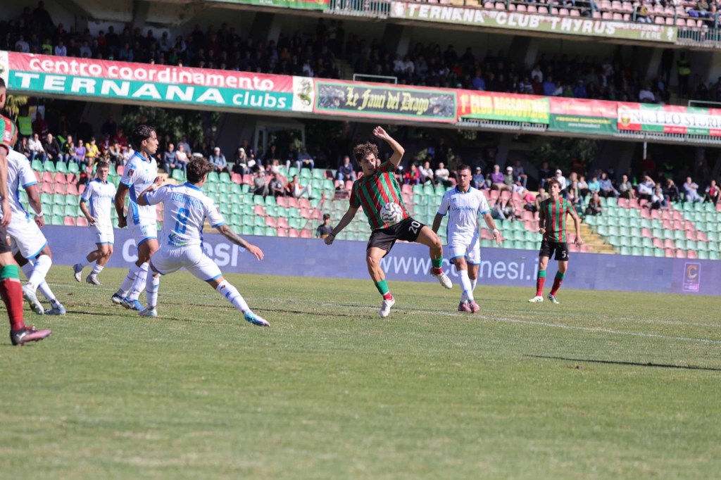 Un giocatore della Ternana in azione durante una partita di calcio, circondato da avversari in maglia bianca, con spettatori sugli spalti.