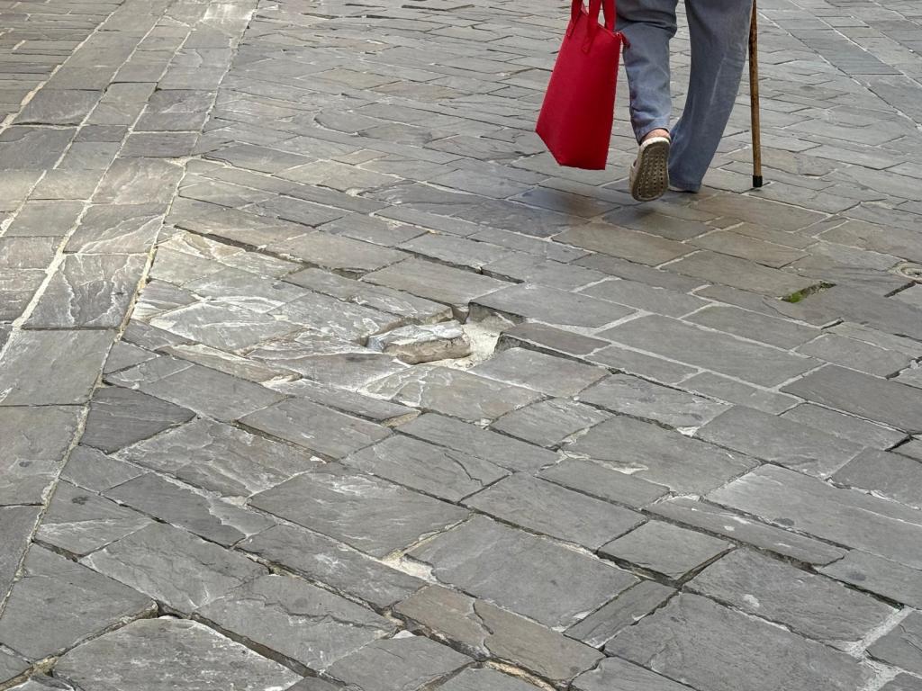 A person walks on a cobblestone street, carrying a red bag and using a cane for support.