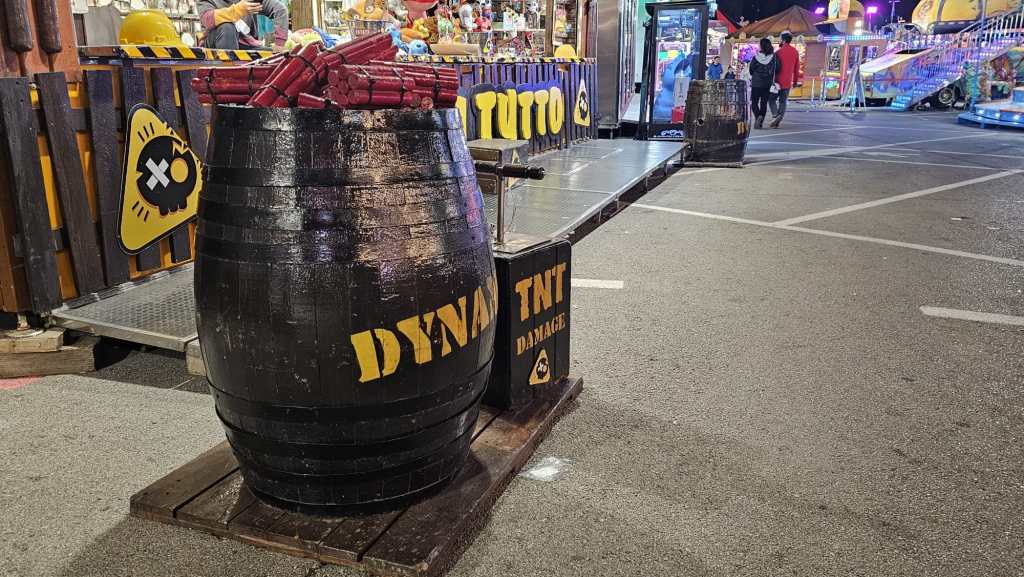 Barrel labeled 'DYNAMITE' filled with red sticks resembling firecrackers at a fairground.