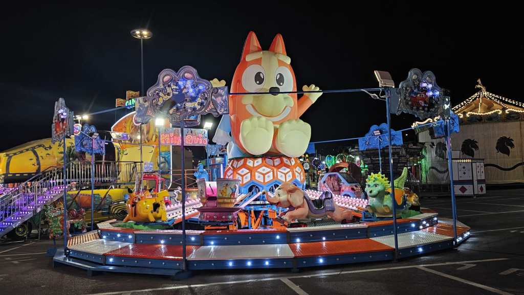 Giostra colorata con un grande animale sorridente al centro, circondata da luci e altre attrazioni da luna park, di notte.