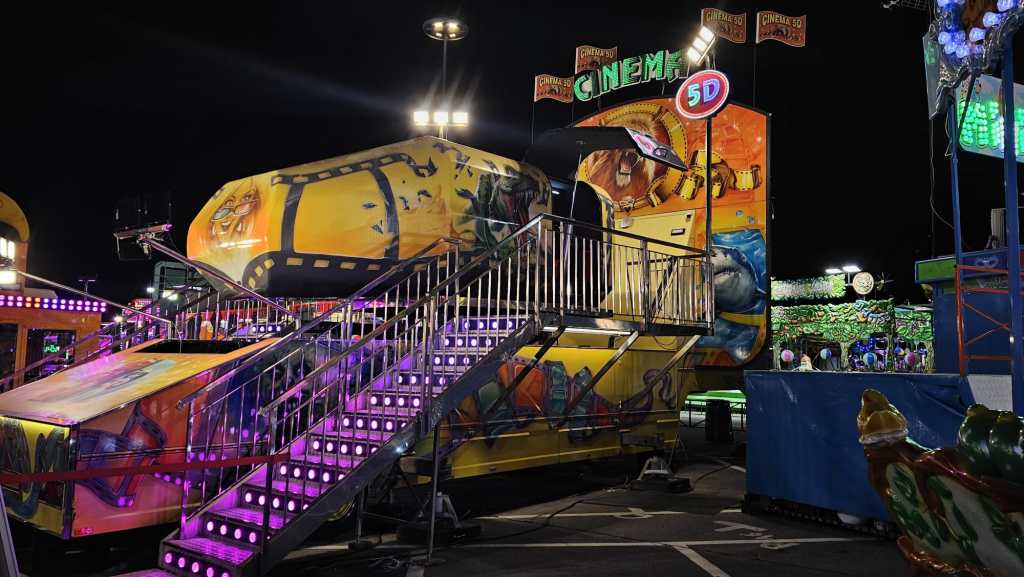 Attrazione di un luna park con un edificio giallo che promuove un'esperienza di cinema 5D, illuminato da luci colorate, in un contesto notturno.