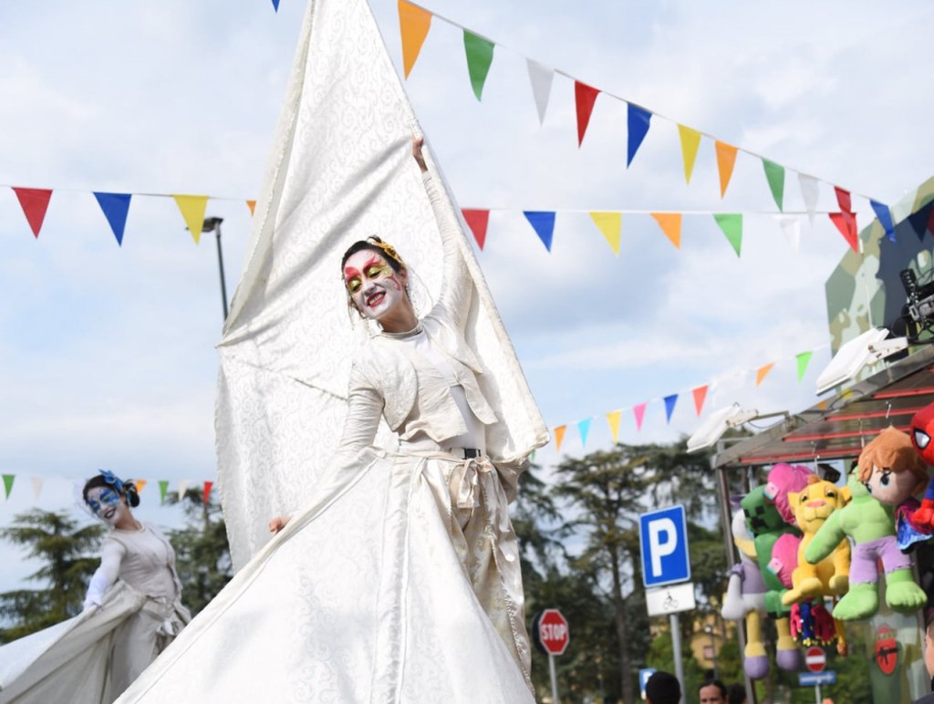 Un'attrice in costume bianco con viso dipinto che danza, circondata da bandierine colorate e figure di peluche appese a un chiosco.