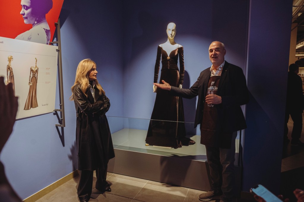 Un uomo e una donna in una galleria d'arte discutono davanti a un manichino che indossa un vestito elegante, con opere d'arte alle pareti in un ambiente moderno.