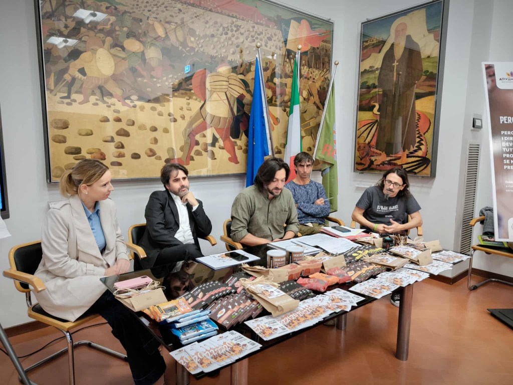 Un gruppo di persone sedute attorno a un tavolo durante una conferenza, con vari materiali pubblicitari e cioccolato artigianale in esposizione, e dipinti sullo sfondo.