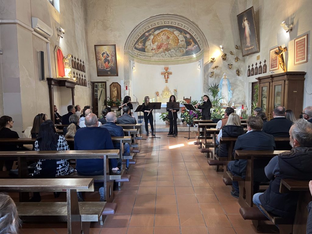 Cerimonia di inaugurazione del restauro della tomba di Giulio Briccialdi, con esibizione musicale in una chiesa. Il pubblico è seduto e ascolta le musiciste, con un ambiente decorato e fiori freschi.