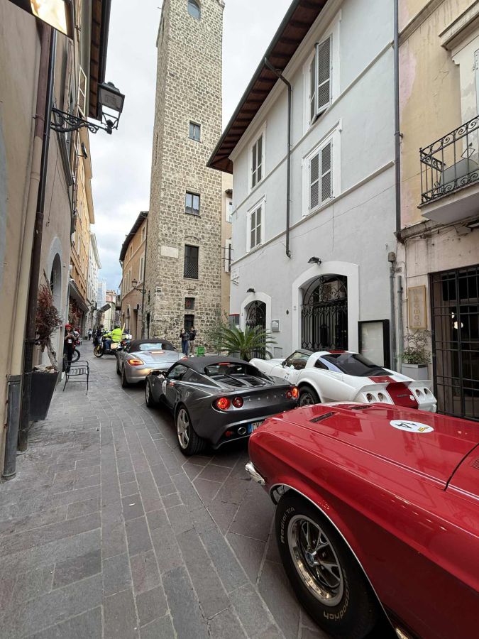 Una strada di via Roma con diverse auto d'epoca e supercar parcheggiate lungo il marciapiede, con un edificio storico sullo sfondo.