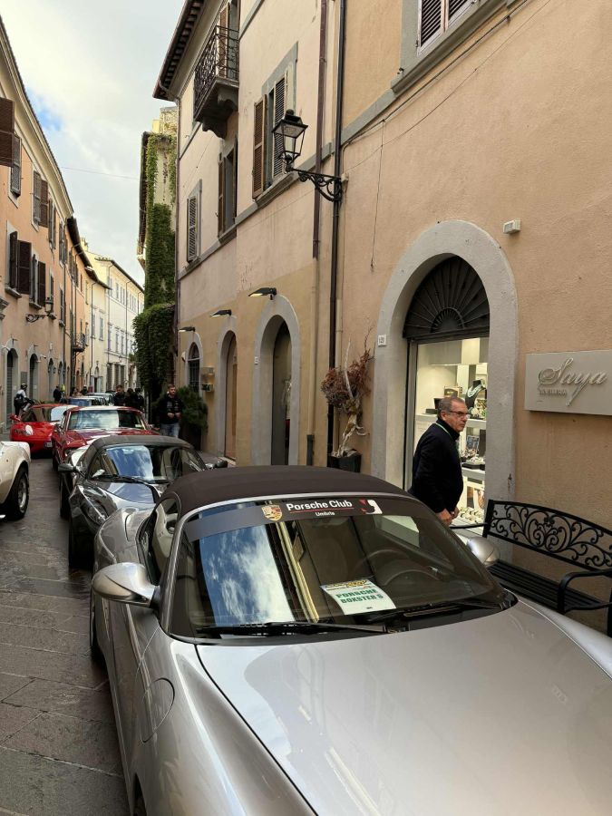 Una strada con auto d'epoca e supercar parcheggiate, circondata da edifici storici. Un uomo in giacca nera cammina accanto alle auto, mentre il cielo è parzialmente nuvoloso.